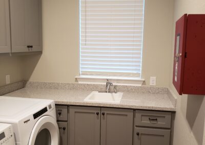 laundry room with white front loader washer and dryer combo with dark red fire extinguisher box on beige walls and gray cabinetry with light gray countertop containing a small wash sink beneath one winder