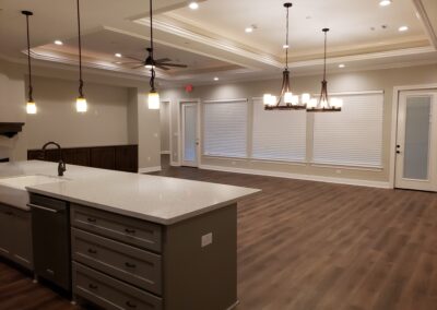 side corner view looking at living room cream fireplace and dark wood mantel from behind gray and white quartz kitchen island with view of dining area's inlet lighted ceiling with double hanging chandeliers