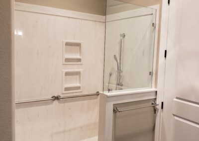 corner view of white marble shower with silver removable shower head and stability handles with glass half wall