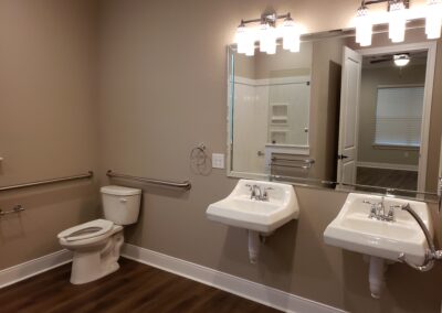 double wheelchair accessible sinks with mirrors and light fixtures above next to toilet area with stability handles with beige colored walls and hardwood flooring