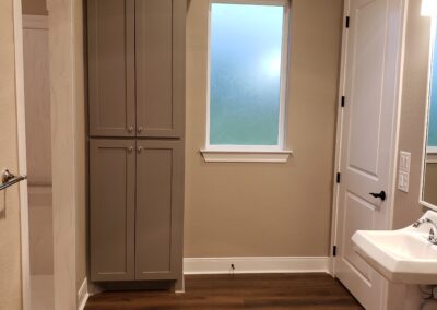 single wall-mounted accessible sink with mirror and light fixture next to door with gray built-in storage cabinet and entry to accessible shower alcove on adjacent wall