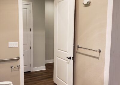 toilet with stability handles next to open door to hallways with entry to accessible shower alcove on the adjacent wall