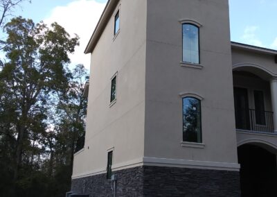 exterior view of narrow three story tan stucco tower attached to the main house and a strip of dark brown stone siding at the bottom